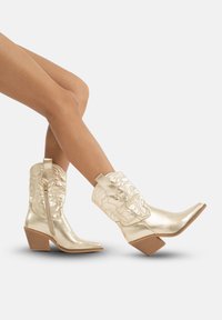 Złote metaliczne kowbojskie buty z pointed toes, wytłaczanym wzorem, bocznym zamkiem błyskawicznym i drewnianym blokowym obcasem. Gładka, błyszcząca faktura na całej powierzchni.