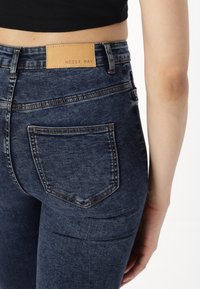 Dark blue denim jeans with a fitted design, featuring a stitched back pocket and a tan leather label reading "NOISY MAY" at the waistband.