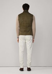 Olivově zelená bezrukávová vesta s prošívaným designem, nošená přes světle hnědý svetr. Doplnená bílými kalhotami a tmavě hnědými botami.