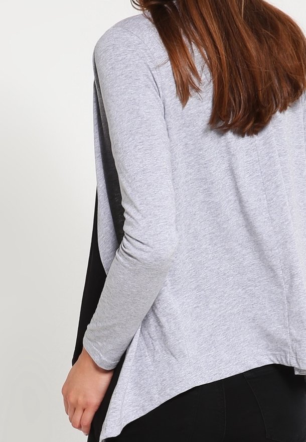 Haut gris à manches longues avec des panneaux latéraux noirs, présentant un ourlet incurvé et une texture douce. Contraste notable entre les matériaux.