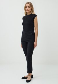 Schwarzes, eng anliegendes Oberteil mit kurzen Ärmeln, kombiniert mit hochtaillierten schwarzen Hosen mit seitlichen Taschen. Das Model trägt schwarze Flats und steht vor einem neutralen Hintergrund.