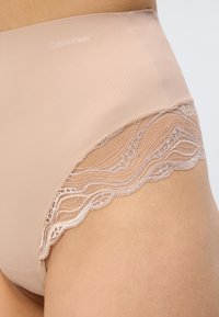 Γυμνό εσώρουχο Calvin Klein με ψηλή μέση και δαντέλα, φορεμένο στο ισχίο και το πάνω μέρος του μηρού ενός ατόμου, πάνω σε ανοιχτό φόντο.