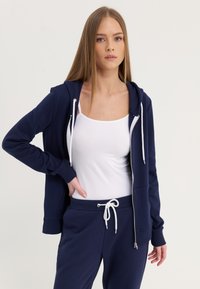 Mornarica zip-up dukserica s bijelim vezicama i džepovima, nosi se preko bijelog topa, u kombinaciji s mornarskim trenirkama s vezicom.