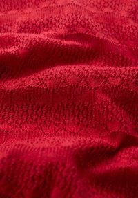 Roter gestrickter Stoff mit kunstvollen Spitzenmustern und wechselnden strukturierten Bereichen, die ein geschichtetes Erscheinungsbild erzeugen.