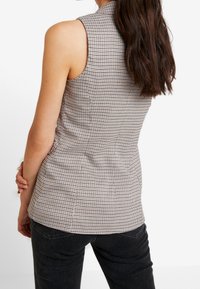 Tajta ärmlös topp i svart och beige hundtandsmönster, med hög halsringning och slät textur. Kombinerad med mörka jeans.