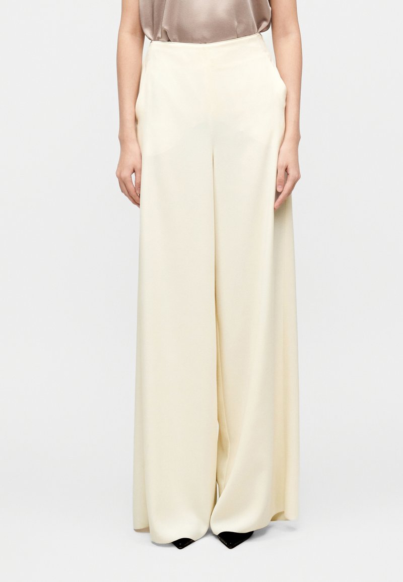 Παντελόνι με φαρδιά πόδια από ανοιχτό κρεμ ύφασμα, με λεία υφή και πλαϊνές τσέπες. Συνδυασμένο με μεταξωτό μπλουζάκι για αντίθεση.