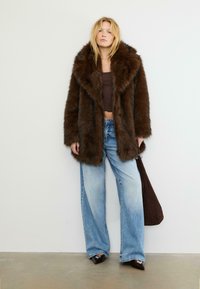 Cappotto in finta pelliccia marrone con ampio colletto, abbinato a jeans a gamba larga blu chiaro. Il modello tiene una borsa marrone scuro, indossando scarpe marroni a punta.