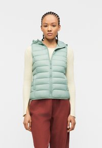 Νέα γυναίκα με πλεξουδάκια στα μαλλιά, φορώντας ανοιχτοπράσινο γιλέκο χωρίς μανίκια με καπιτονέ σχέδιο, ριμπ καφέ μακρυμάνικη μπλούζα και παντελόνι σε σκουρόχρωμή σκουριά, όρθια.