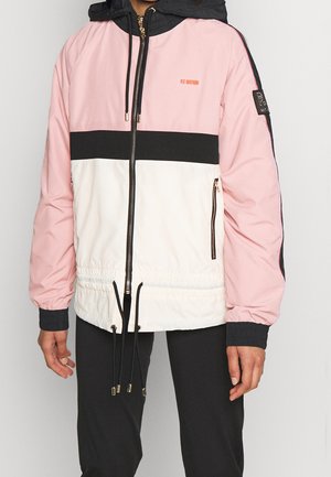 Osoba ubrana w różowo-czarno-białą kurtkę typu zip-up z kolorowymi blokami, z troczkami i kieszeniami na zamek, w połączeniu z czarnymi spodniami.