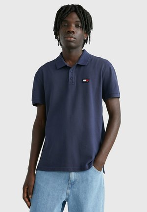 Camisa polo de algodón azul marino con una tapeta de tres botones, mangas cortas y un pequeño parche de logo en el pecho izquierdo. Llevada con jeans de color azul claro.