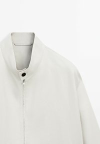 Helle graue Jacke mit Stehkragen, ausgestattet mit einem Reißverschluss vorne und einem schwarzen Knopf. Hergestellt aus glattem, strukturiertem Stoff mit minimalistischer Gestaltung.