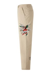 Pantalon beige avec poches latérales présentant une broderie colorée d’un oiseau, d’un cœur, d’un poignard et une bannière avec l’inscription "TRUE TILL DEATH."