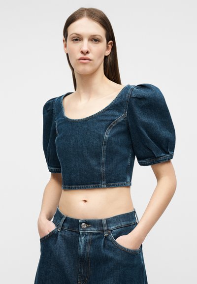 Γυναίκα με σκουρόχρωμο μπλε cropped denim τοπ με φουσκωτά μανίκια και αντίστοιχο τζιν παντελόνι ψηλόμεσο, με τα χέρια στις τσέπες.