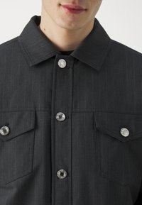 Svart randig skjortjacka med klassisk krage, silverfärgade tryckknappar och två bröstfickor med klaff.