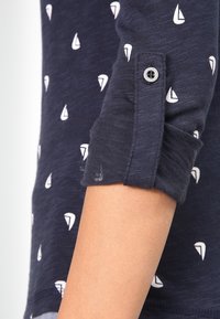 Chemise à manches longues bleu marine avec un motif de voilier blanc, dotée d'un esprit retroussé et d'un détail à bouton unique près de la manche.