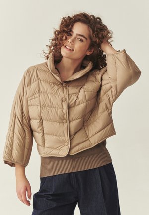 Mujer joven con cabello rizado que lleva una chaqueta corta acolchada beige y pantalones oscuros, sonriendo con una mano tocándose la cabeza.