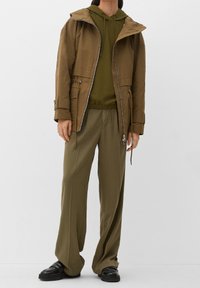 Chaqueta de capucha verde oliva con cremallera en el frente y cordón, combinada con un suéter texturizado verde a juego y pantalones holgados, llevados con zapatos negros.