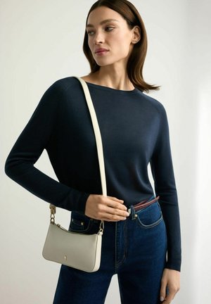 Kobieta w granatowym swetrze i ciemnych dżinsach, niosąca małą beżową torebkę na ramię ze złotymi okuciami.