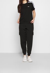 Schwarzes T-Shirt mit kurzen Ärmeln, weißen Schulterstreifen und Logo auf der Brust, kombiniert mit schwarzen Sweatpants mit Bündchen und seitlichen Cargo-Taschen.