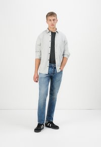 Camisa gris claro, camiseta negra, jeans azules y zapatillas negras con acentos blancos. El modelo está de pie contra un fondo blanco.