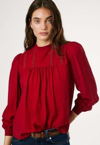 Blusa rossa a maniche lunghe con collo alto, tessuto testurizzato, dettagli arricciati e accenti sottili, indossata sopra jeans scuri e una cintura marrone.