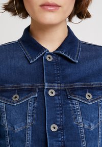 Mörk denimjacka med klassisk krage, knappar i silverton, två bröstfickor och kontrasterande sömnadsdetaljer.