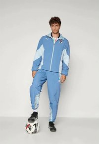 Fato de treino atlético azul feito de material leve, com uma jaqueta com fecho éclair e detalhes coloridos, e calças a condizer, acompanhado de uma bola de futebol.