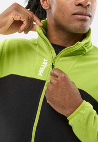 Green and black fleece zip-up jacket with ribbed texture. Features a high collar and zipper, with a logo on the upper left.