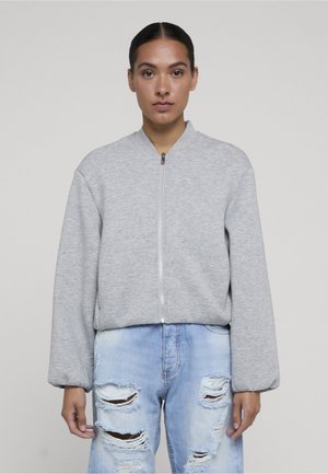 Femme aux cheveux foncés portant une veste grise zippée et un jean bleu clair déchiré, debout devant un fond uni.