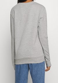 Personne portant un sweat-shirt à manches longues gris clair uni et un jean bleu ample, vue de dos sur un fond uni.