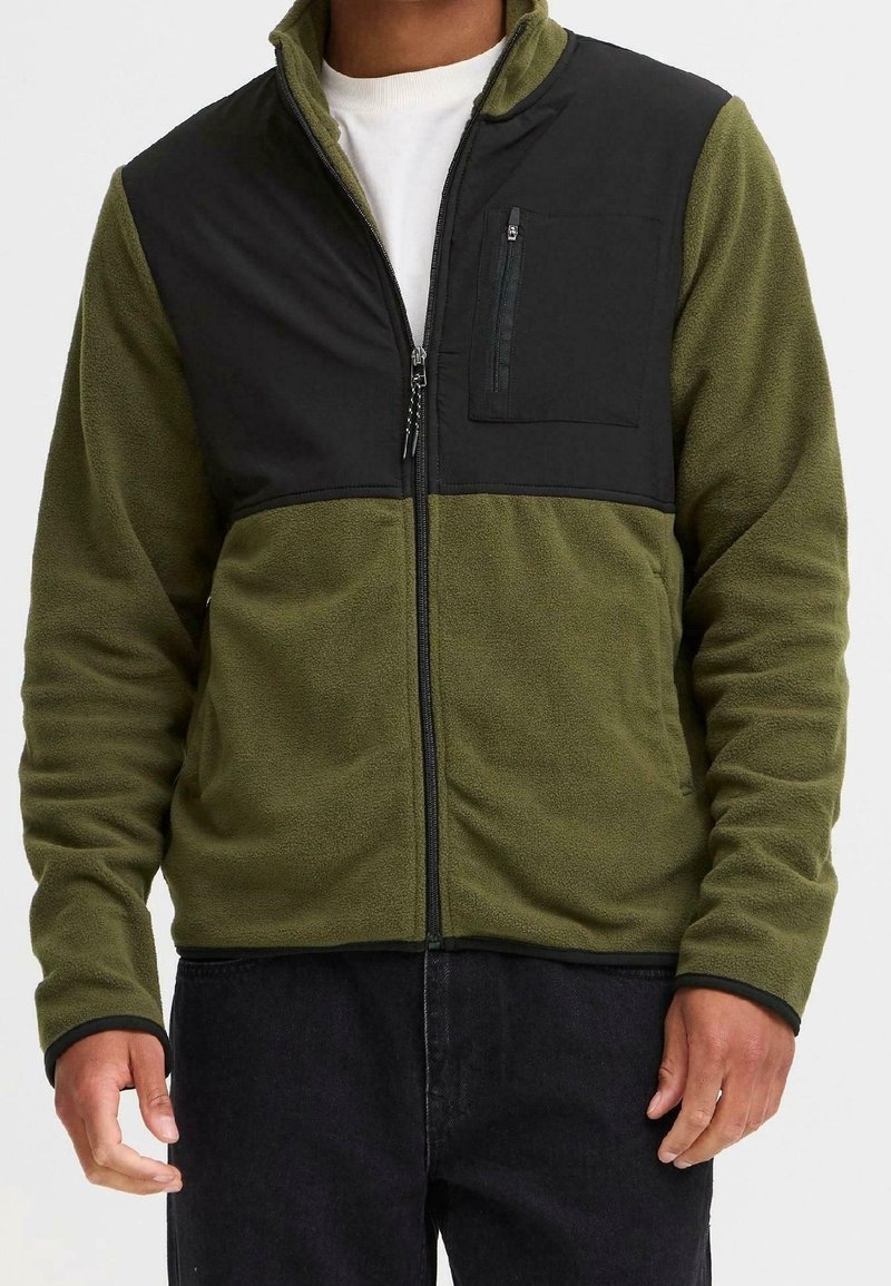 Homme portant une veste polaire verte avec un panneau noir sur la partie supérieure de la poitrine et une poche zippée, chemise blanche en dessous et pantalon noir.