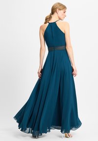 Langes, fließendes Teal-Kleid mit Neckholder-Ausschnitt, strukturiertem Taillenbund und glattem Stoff. Sanfte Falten betonen Bewegung und Drapierung.