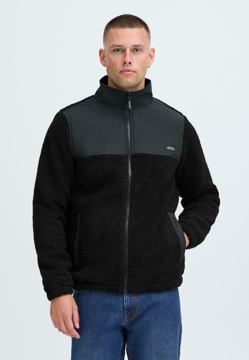 Giacca in fleece nera con un pannello scuro nella parte superiore, zip completa, colletto alto e due tasche laterali. Texture morbida e vestibilità rilassata.