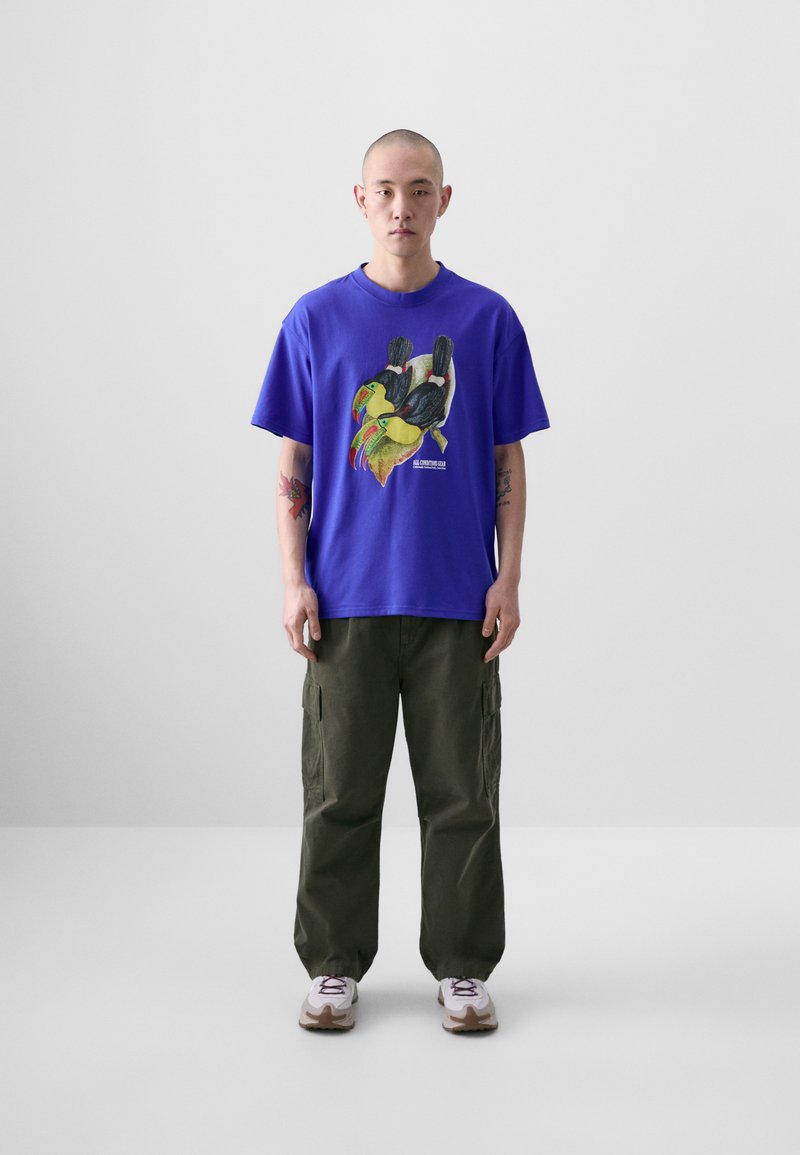 T-shirt en coton violet avec un graphique coloré de toucan, associé à un pantalon cargo vert olive et des baskets blanches. Design simple et décontracté.