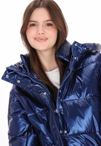 Csillogó sötétkék steppelt dzseki magas gallérral, cipzáras zárással és patentgombokkal. Steppelt részekkel és sima, tükröződő felülettel rendelkezik.