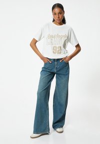 Camiseta blanca con gráfico "Los Angeles League 92" en dorado, combinada con jeans de mezclilla anchos de color azul y zapatillas blancas, con un ajuste relajado.