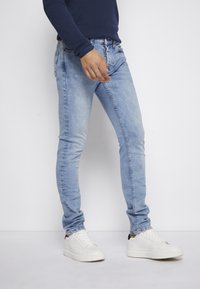 Jeans skinny biru muda terbuat dari denim, dengan desain lima saku klasik. Dipadukan dengan sepatu sneakers putih dan atasan lengan panjang navy.