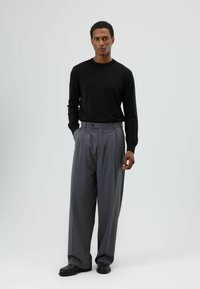 Haut noir à manches longues, pantalon gris plissé avec détail de boutons, porté avec des chaussures noires, mettant en avant une coupe structurée et un tissu lisse.