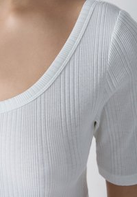 Top bianco a maniche corte con scollatura a V ampia. La texture del tessuto è morbida, leggera e leggermente trasparente con linee verticali.