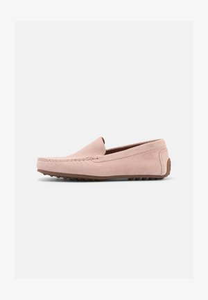 Persoon die lichtroze suède loafers en donkere broek draagt, voeten rustend op een metalen rechthoekig blok tegen een witte achtergrond.