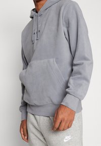 Felpa grigia in pile con tasca frontale, cappuccio con cordino, polsini a costine e logo ricamato "AIR" sul petto. Indossata con pantaloni della tuta grigi.