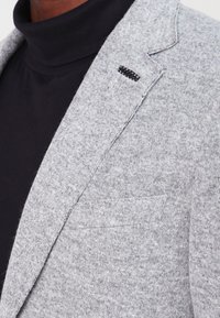 Primer plano de una persona que lleva una chaqueta gris texturizada sobre un suéter de cuello alto negro, enfocándose en la solapa de la chaqueta y el bolsillo del pecho.