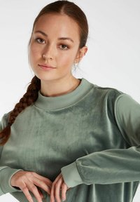 Groene, fluwelen sweatshirt met een geribde kraag en manchetten. De stof heeft een gladde textuur en heeft een loose fit en lange mouwen.