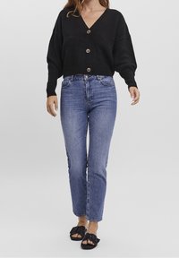 Zwart gebreide cropped cardigan met een v-hals, voorzien van grote knopen, gecombineerd met hooggerokte blauwe jeans en zwarte instapsandalen.