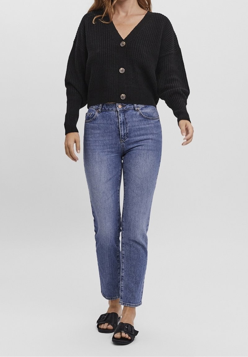 Zwart gebreide cropped cardigan met een v-hals, voorzien van grote knopen, gecombineerd met hooggerokte blauwe jeans en zwarte instapsandalen.
