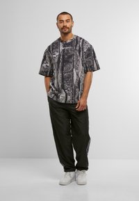 Mand står med hænderne i lommerne iført en mønstret sort og grå oversized t-shirt, sorte bukser og hvide sneakers foran en ensfarvet baggrund.