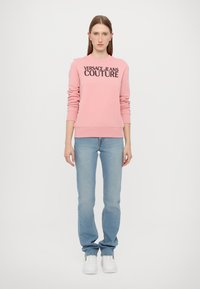 Rozā kokvilnas sporta krekls ar "VERSACE JEANS COUTURE" logotipu, apaļu kakla izgriezumu, ribotām piedurknēm un apakšmalu, savienots ar gaiši ziliem džinsiem un baltiem sporta apaviem.