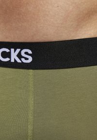 Grüne Stoff-Boxershorts mit einem schwarzen Gummibund, der mit weißem Schriftzug versehen ist. Das Material ist glatt und zeigt sichtbare Nähte.