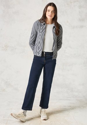Cardigan gris à motifs avec une fermeture éclair, haut gris clair et jean large bleu marine. Chaussures comprenant des baskets grises et blanches. Fond neutre.