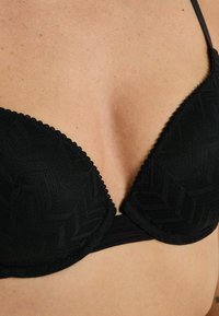 Primo piano di un décolleté che indossa un reggiseno nero texture con spalline sottili e motivi geometrici sul tessuto.